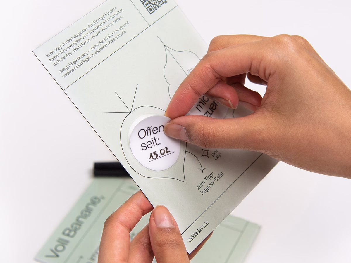 A hand peels a round sticker from the edge of an Odds & Ends postcard.