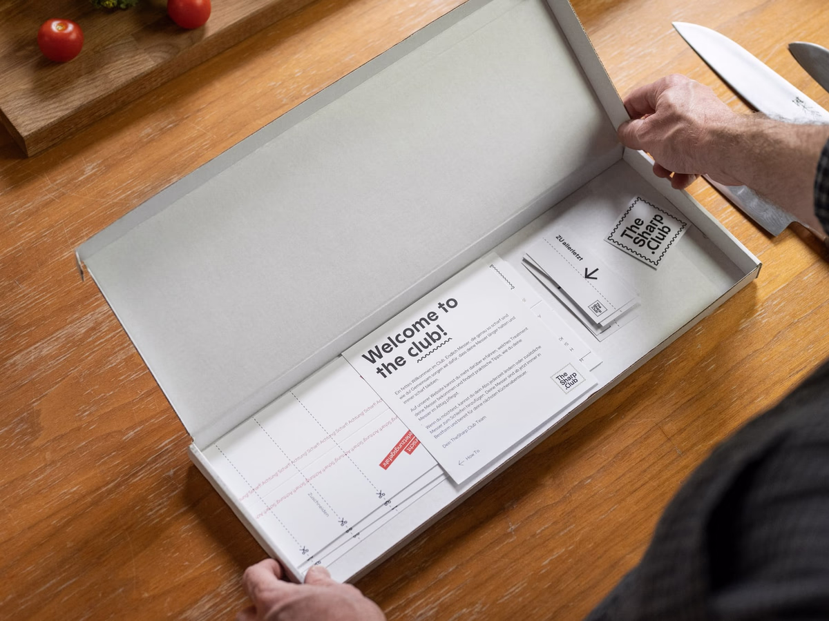 Opened TheSharp.Club box on a wooden table, photographed from above with welcome card and kitchen knife.