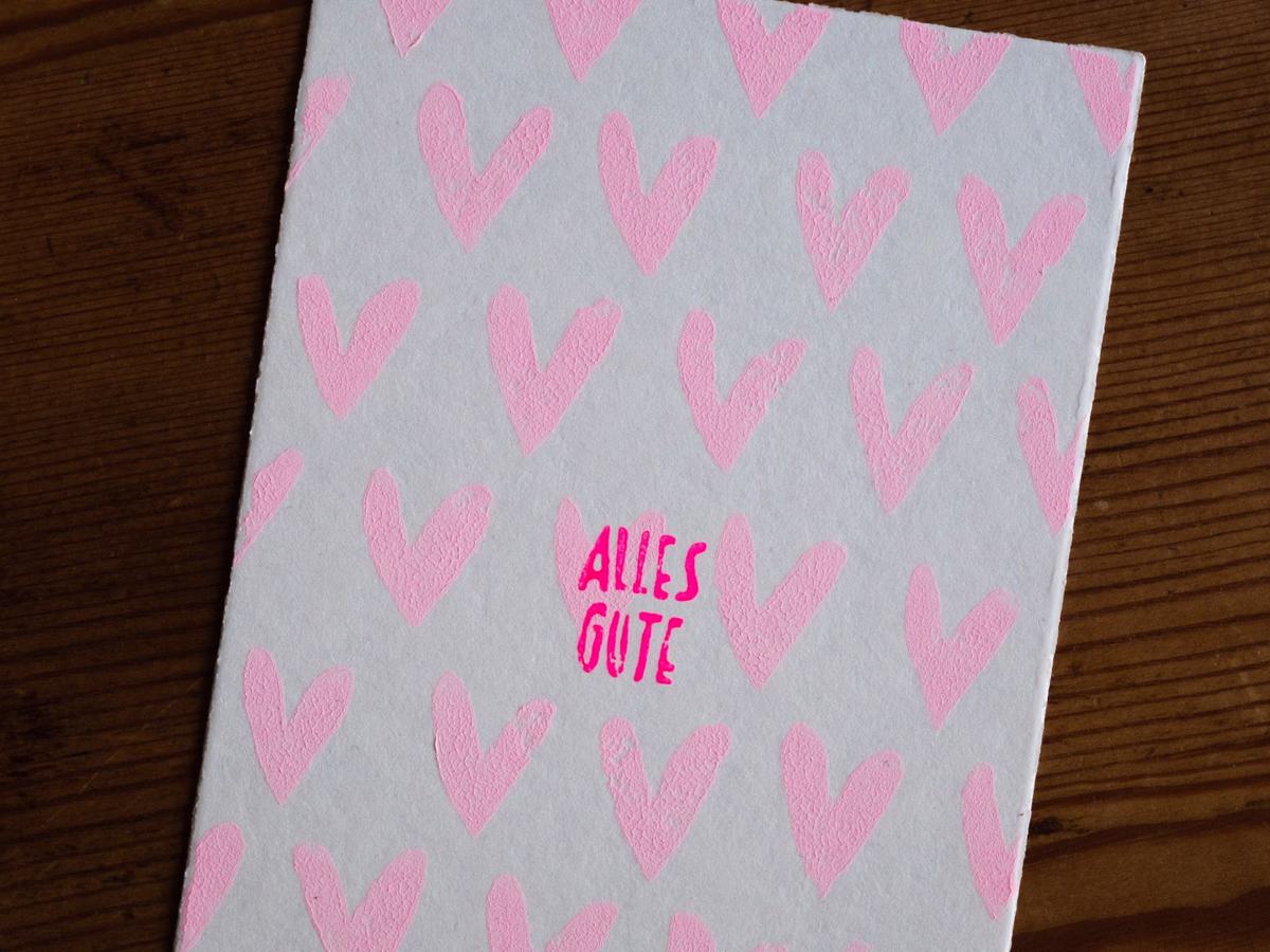 Linocut postcard with neon-pink hearts and the lettering "Alles Gute", leaning against a light wall.
