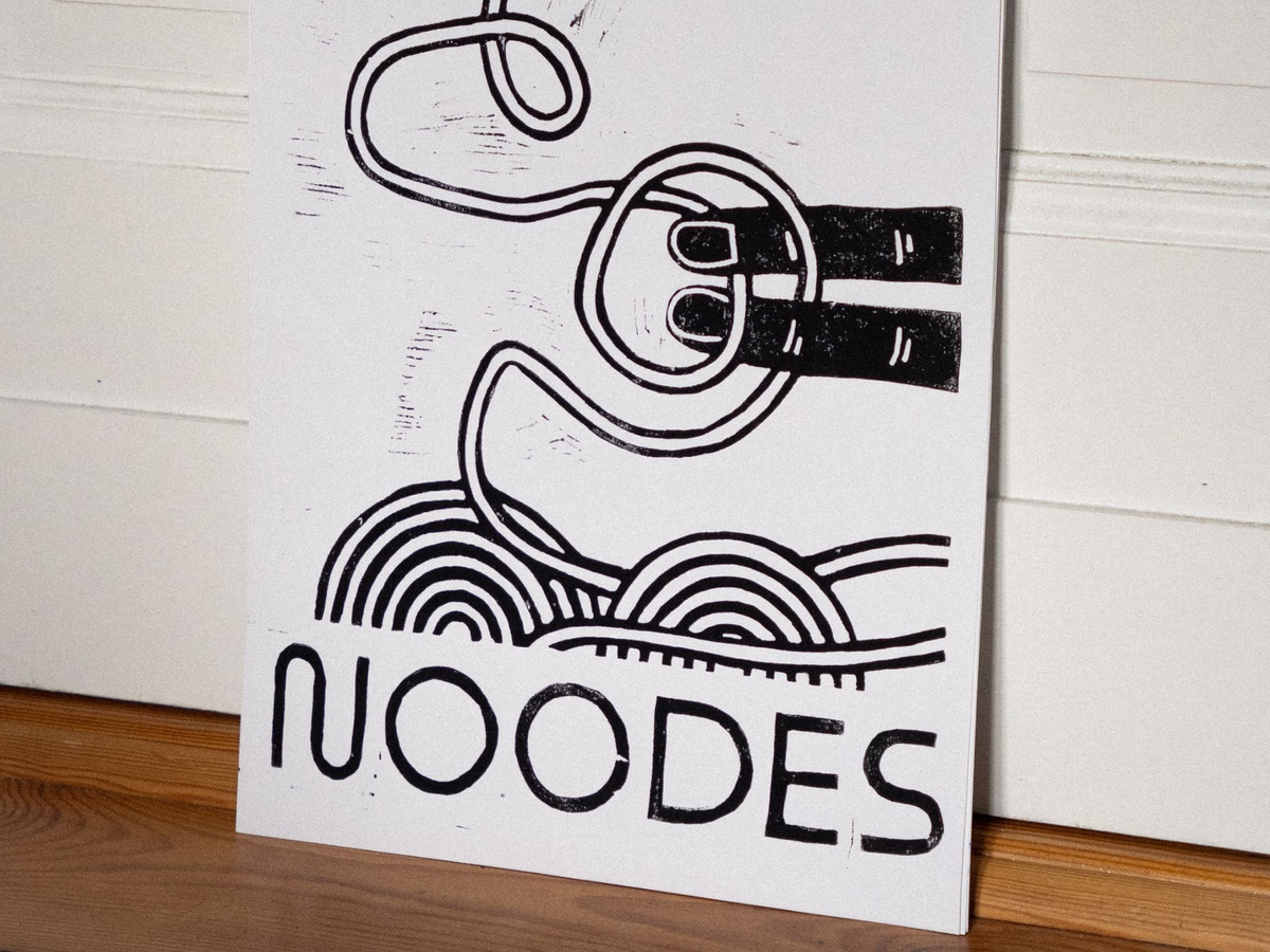 Black and white A3 linocut with noodle motif and lettering "NOODES", leaning against a light wall.