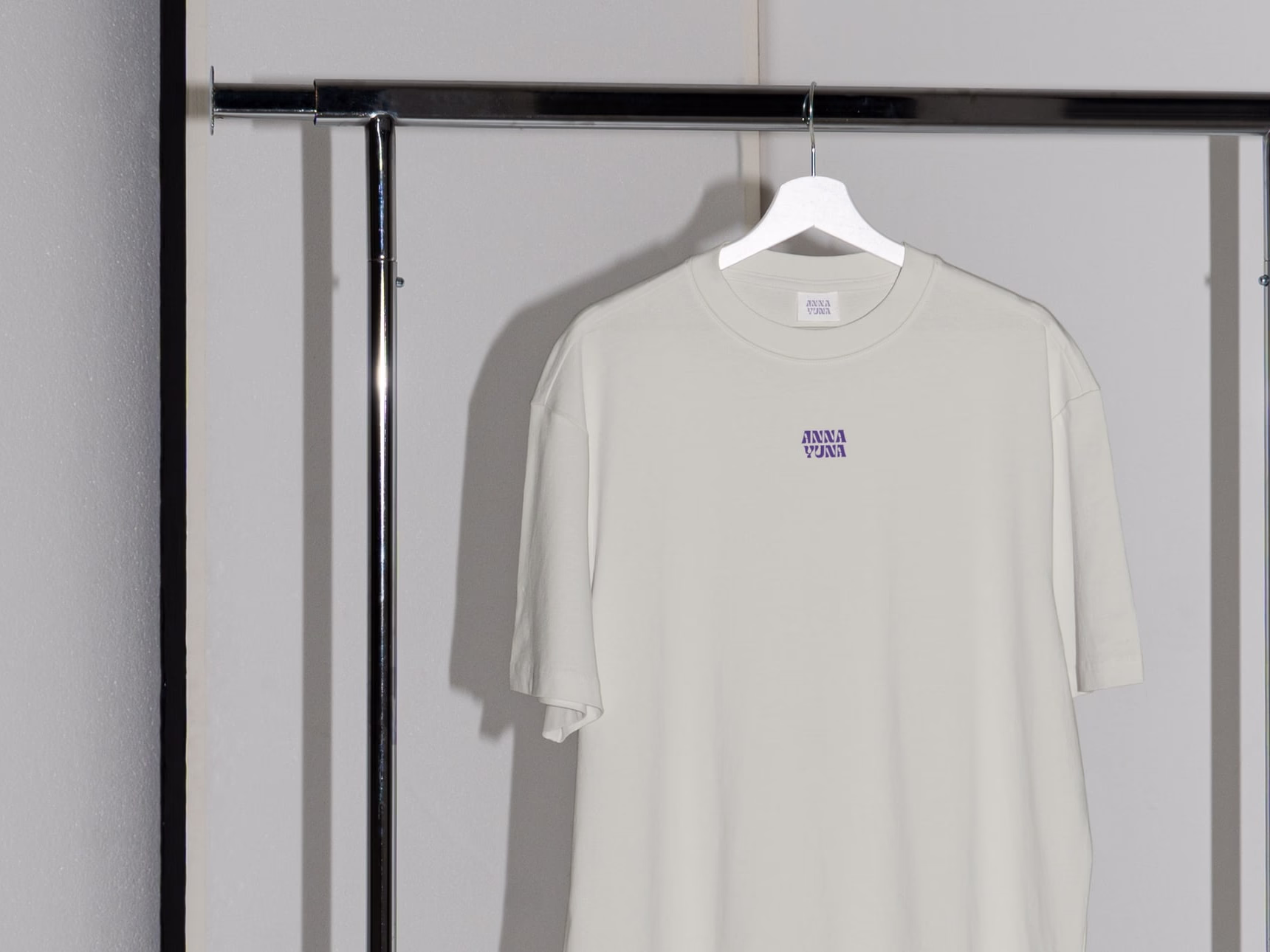 Cream-coloured t-shirt with a small sewn-on ANNA YUNA wordmark patch, hung on a metal rack.