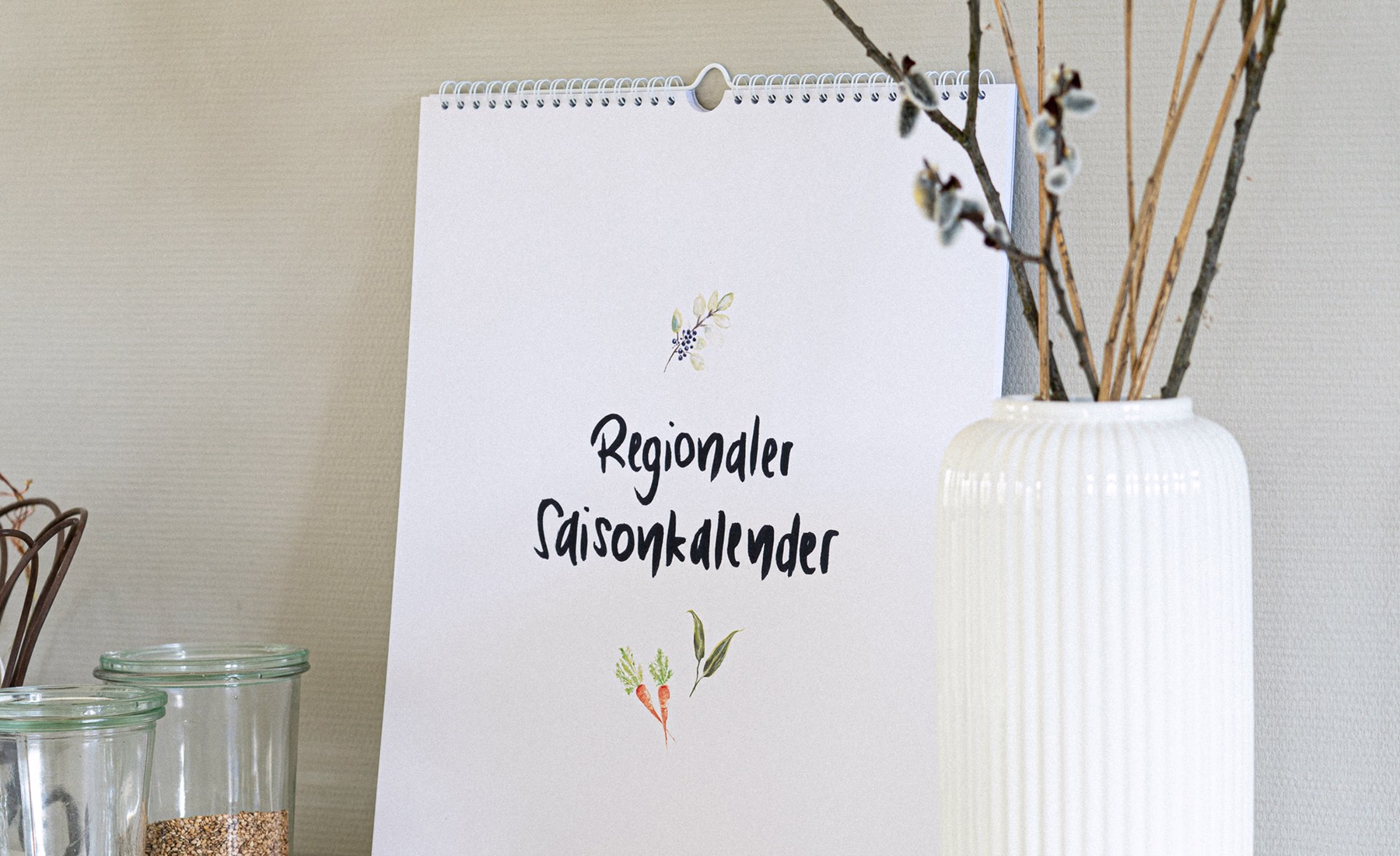 Hand-drawn seasonal calendar of regional vegetables and fruit, hung on a light wall next to a vase.